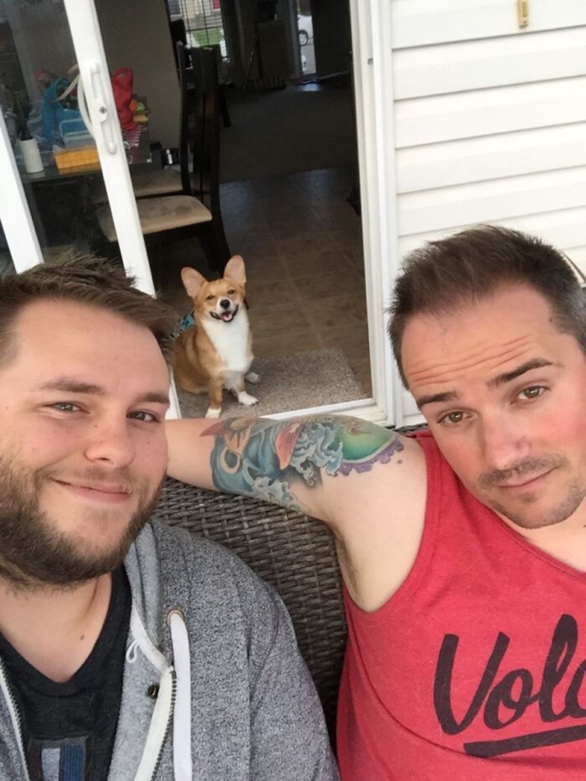 brown and white corgi sitting on the floor behind two men taking a selfie