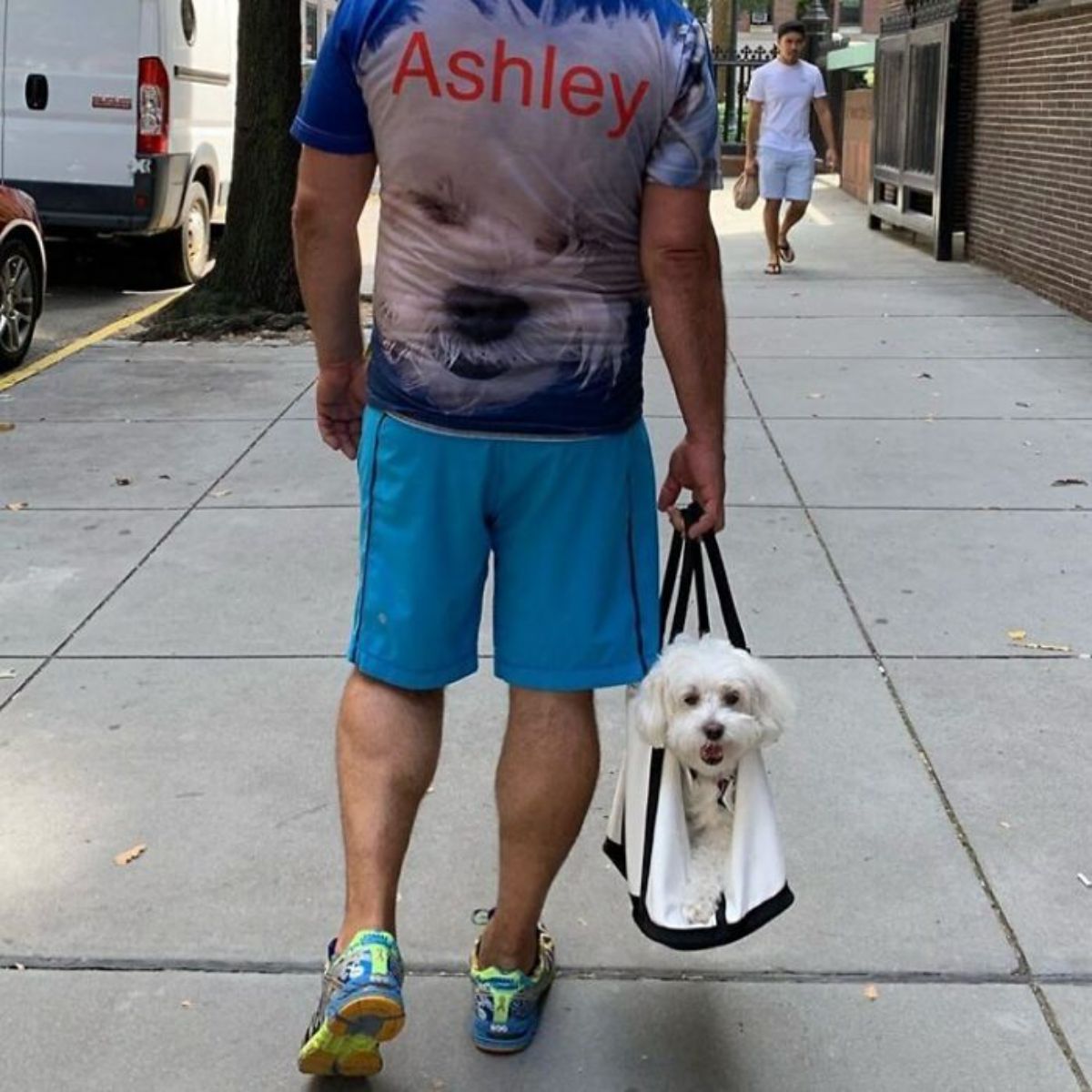 a white fluffy dog in a white and black dog being carried on the road by someone wearing a shirt with the dog's face on it