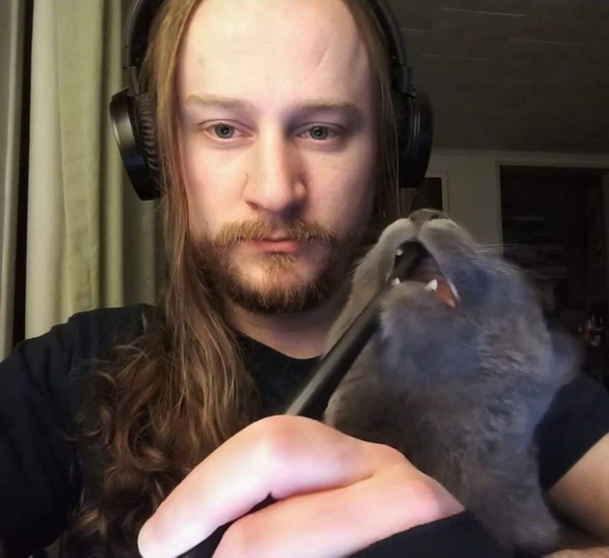 man holding a paint brush with the other end inside the mouth of a grey cat sitting on his lap