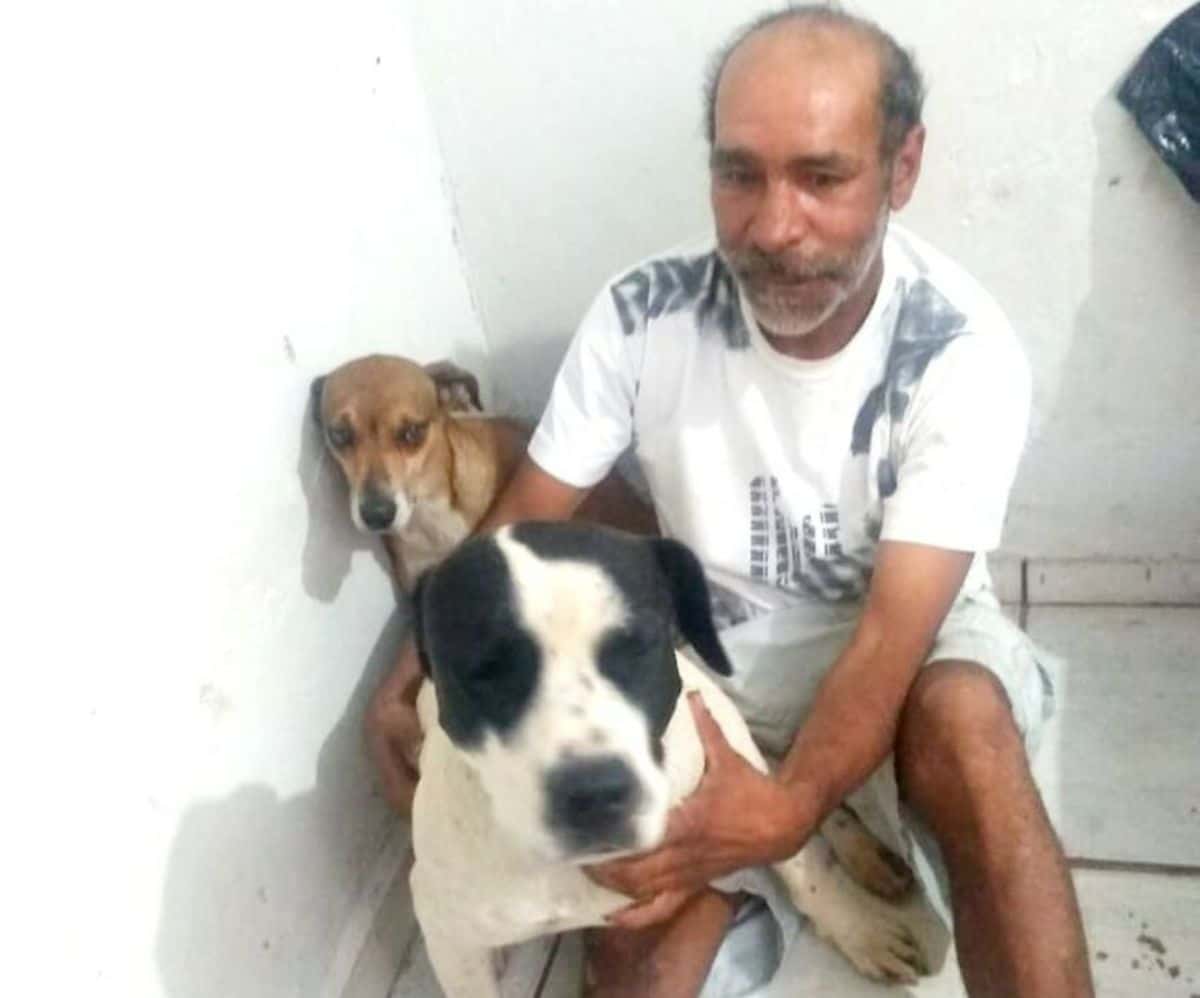 old man sitting on the floor next to a brown and white dog and holding a black and white dog