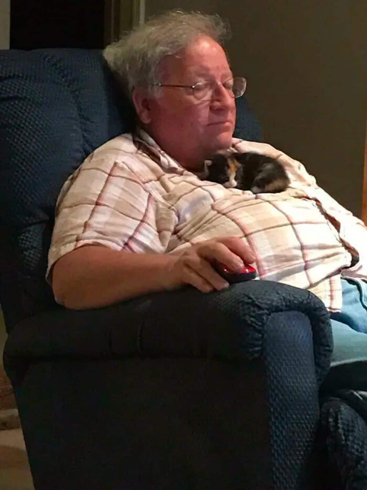orange black and white kitten sleeping on an old man's chest while the man is sitting up on a black chair