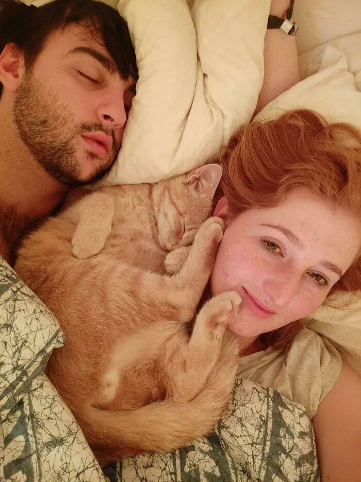 orange cat sleeping on a bed between a couple with the cat's legs and body on the woman