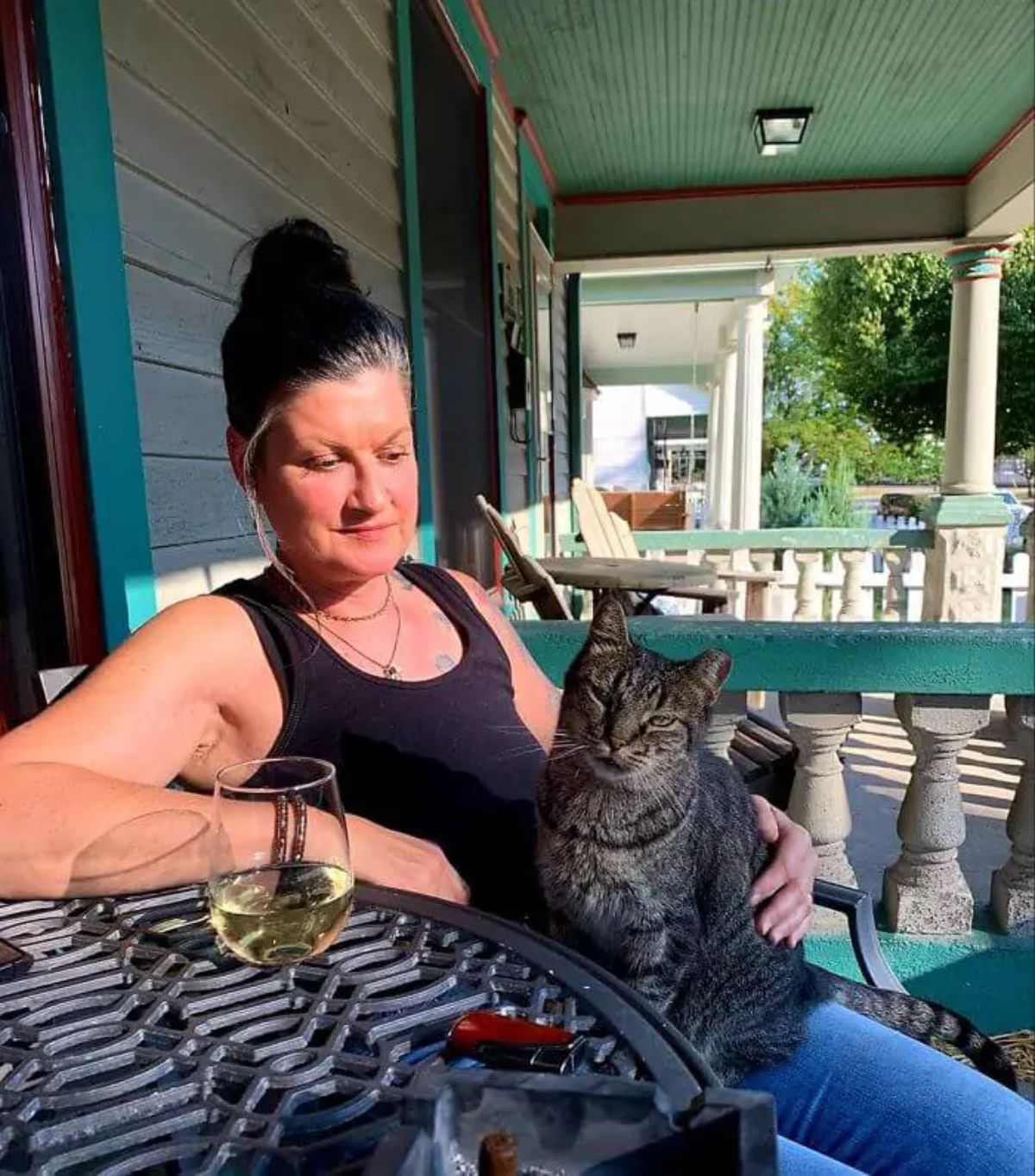 woman sitting outside with an arm on a black patio table with a glass of wine having a grey tabby cat sitting on her lap