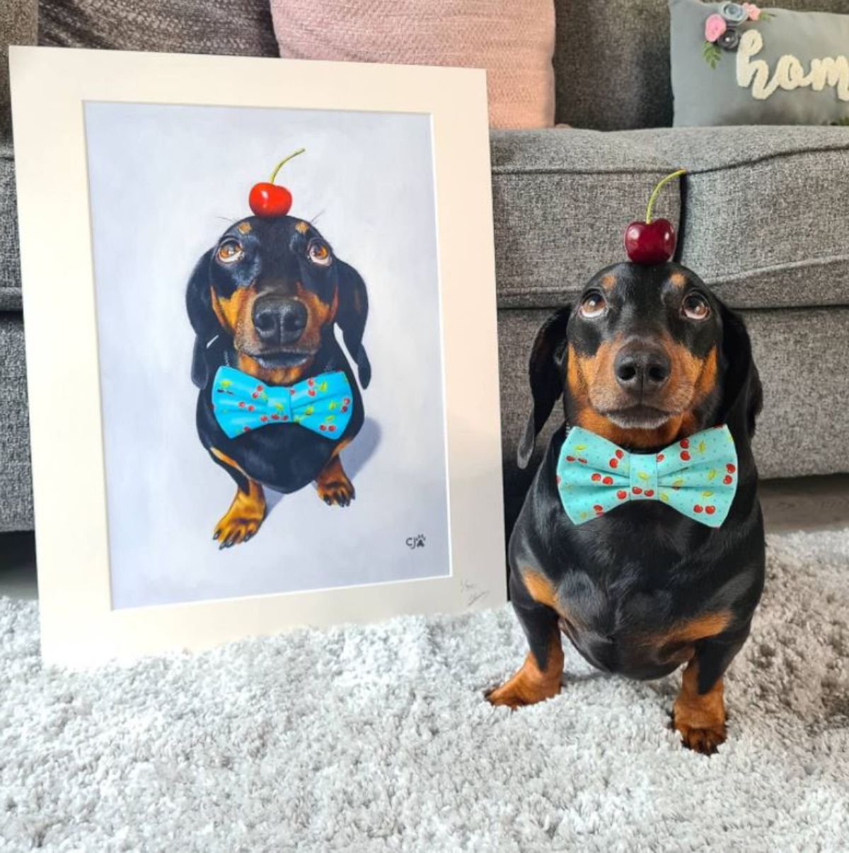 black and brown dachshund with a cherry on the head wearing a blue and red cherry bowtie next to a painting of the dog in the same pose and outfit