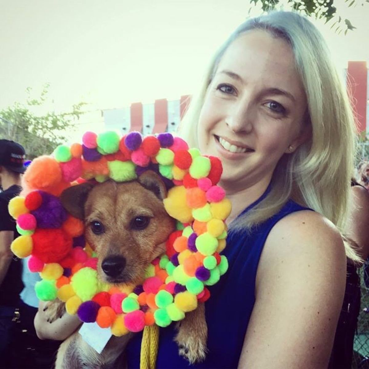 brown dog wearing an elizabethan cone with colourful pompoms on it being held by a smiling woman