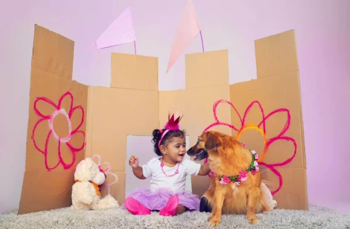 little girl dressed as a princess with a brown dog in flower necklace in front of a cardboard palace