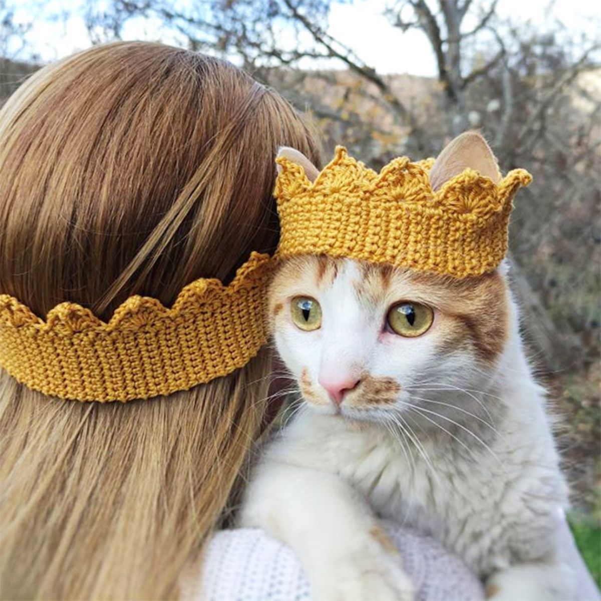 white and orange cat wearing a yellow crocheted crown held by someone wearing a yellow crocheted crown headband