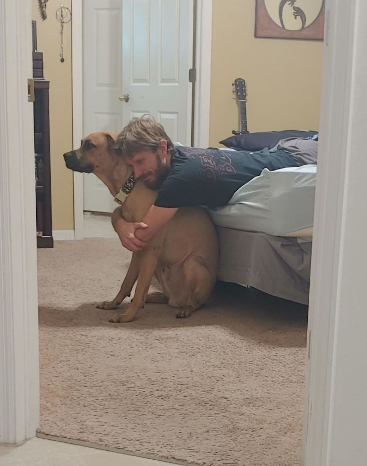 brown dog sitting on floor getting hugged by a man laying on a bed
