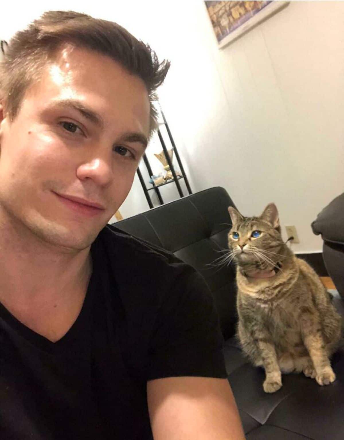 chonky brown tabby cat sitting on a black sofa looking lovingly at a man next to her