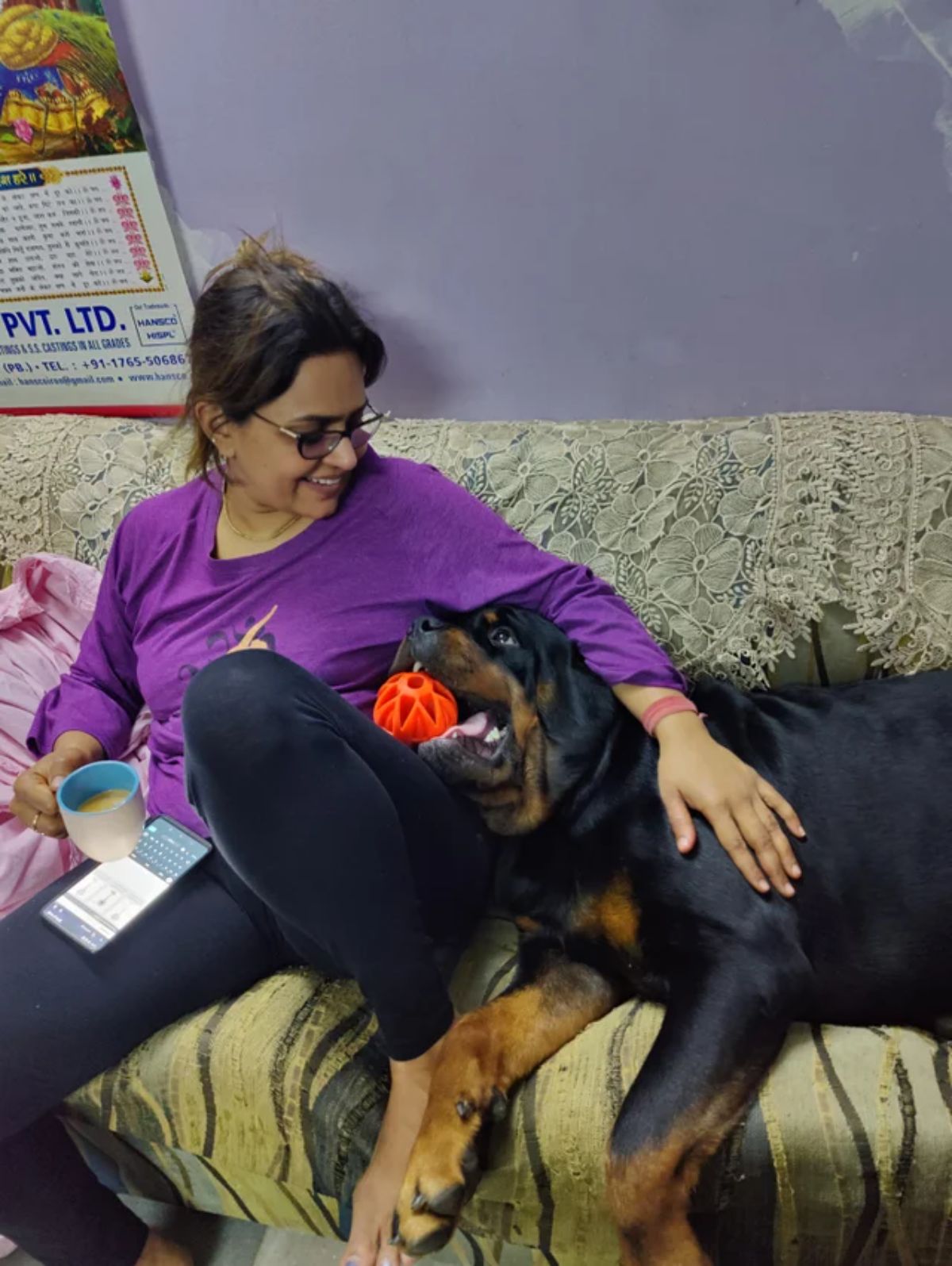 large black and brown dog laying on a sofa with an orange ball in the mouth looking up at a woman lovingly