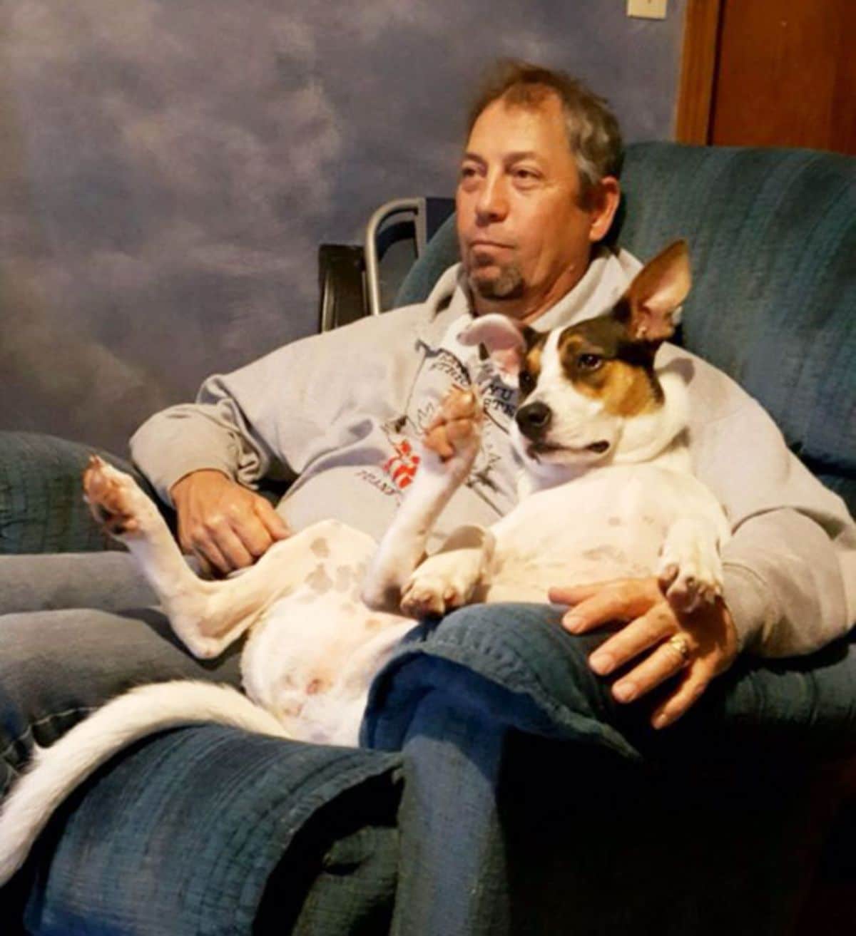 man sitting on green chait with a white and brown dog laying belly up on his lap