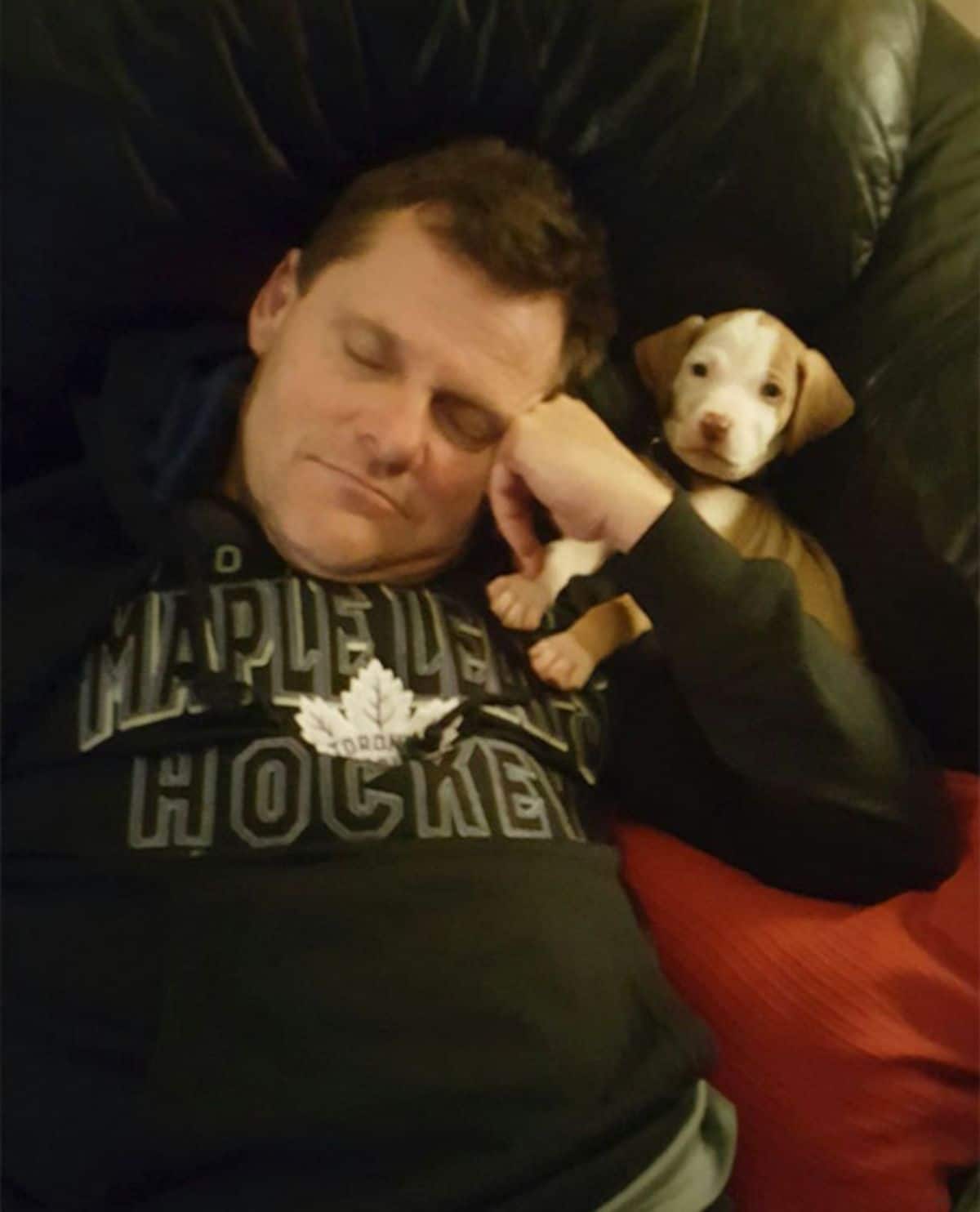 man sleeping with a brown and white puppy on his shoulder
