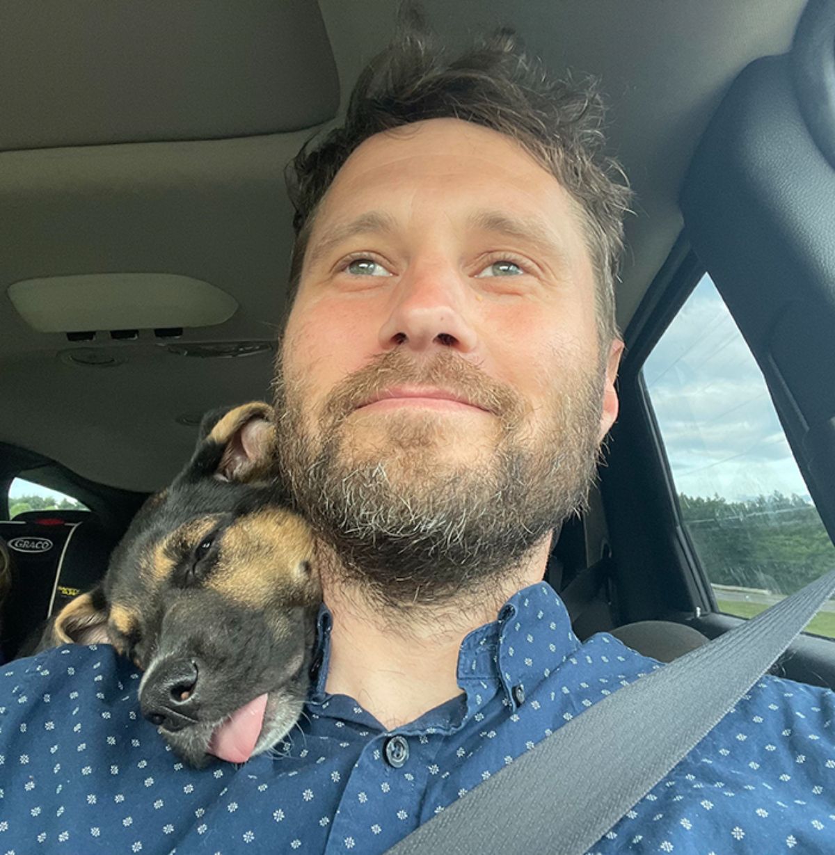 black and brown puppy with the tongue out and head on a man's shoulder while the man's sitting at the driver's seat