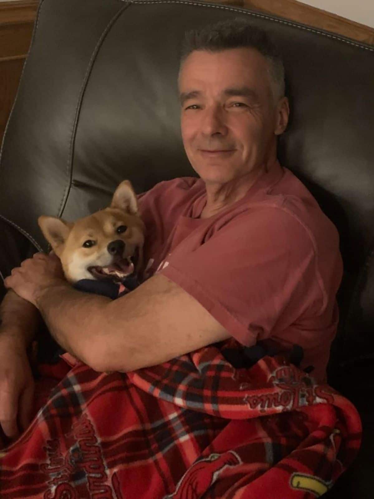 brown and white shiba inu being cuddled by a smiling old man on a black sofa