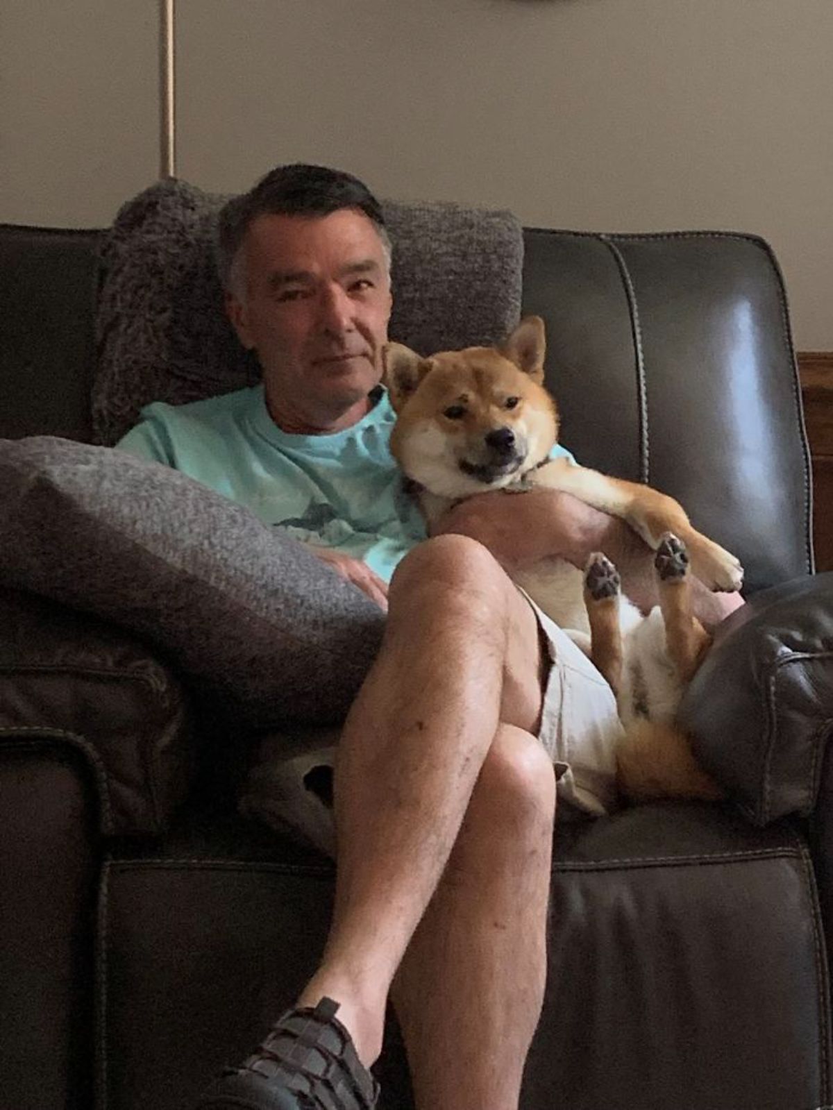 brown and white shiba inu sitting up and being hugged by an old man sitting on a black sofa