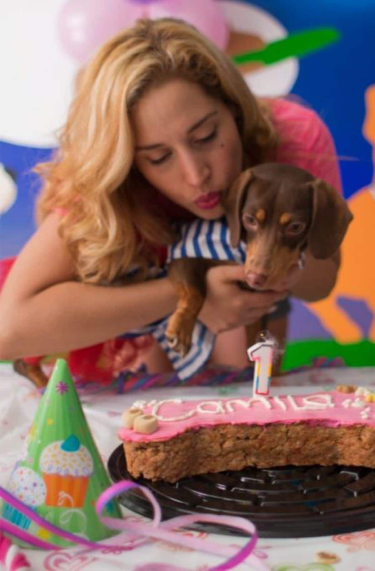 brown dachshund being held by a woman in front of a pink and brown cake with a 1 candle on it