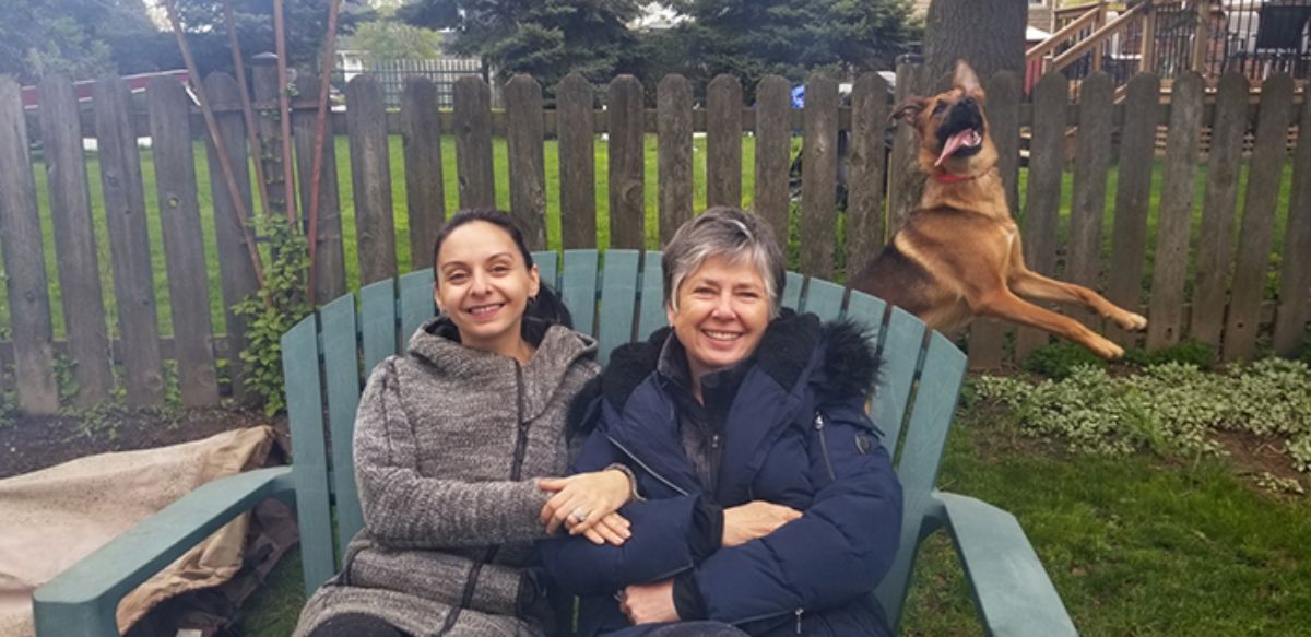 brown dog with tongue out jumping behind a woman and an older woman on a bench