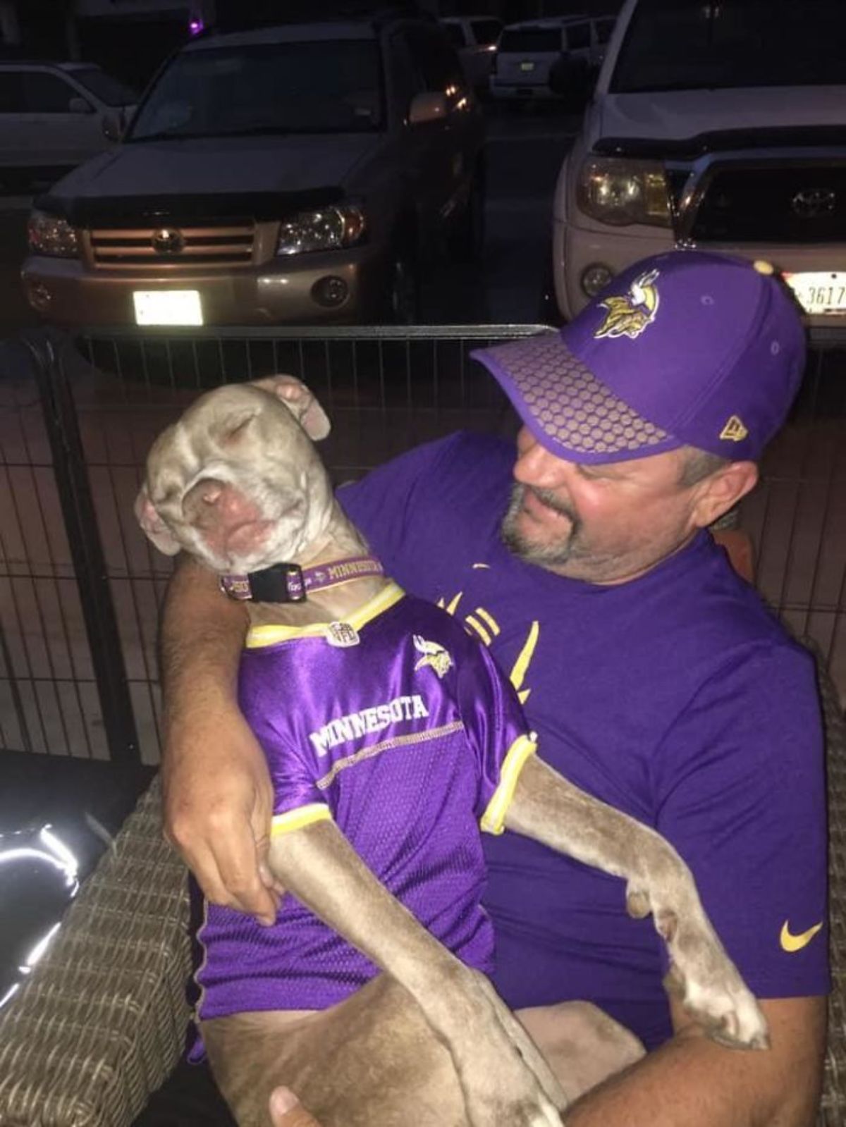 grey and white pitbull sleeping in a man's arms and both are wearing matching purple and yellow shirts
