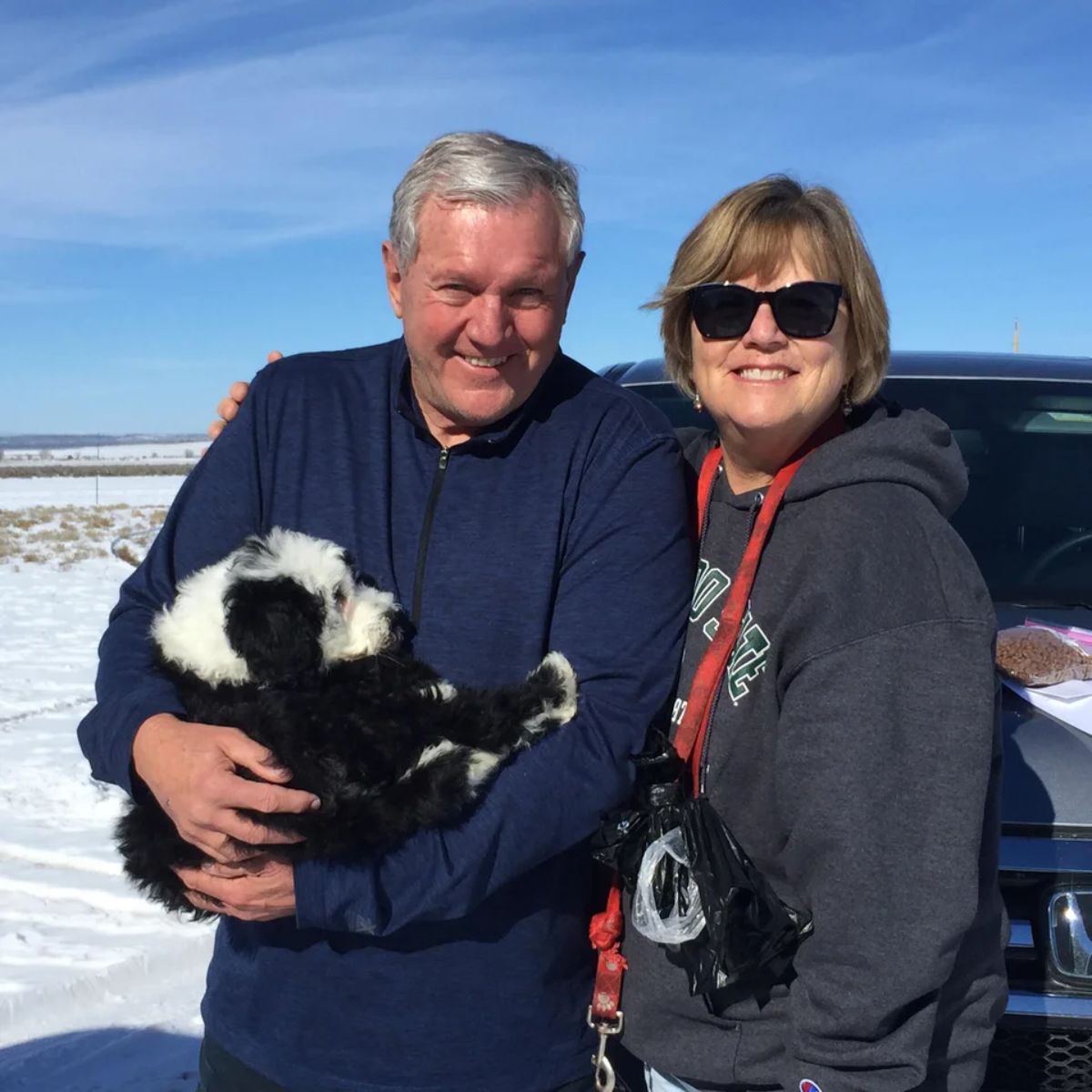 small fluffy black and white puppy being cuddled by an old man who is posing with an old woman in front of a vehicle in a snowy landscape