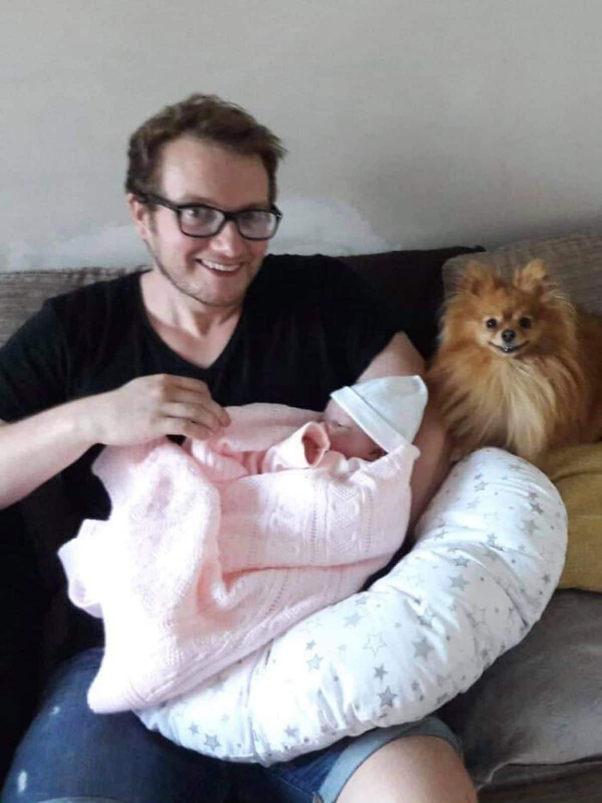 smiling fluffy brown pomeranian sitting next to a man holding a baby in his arms