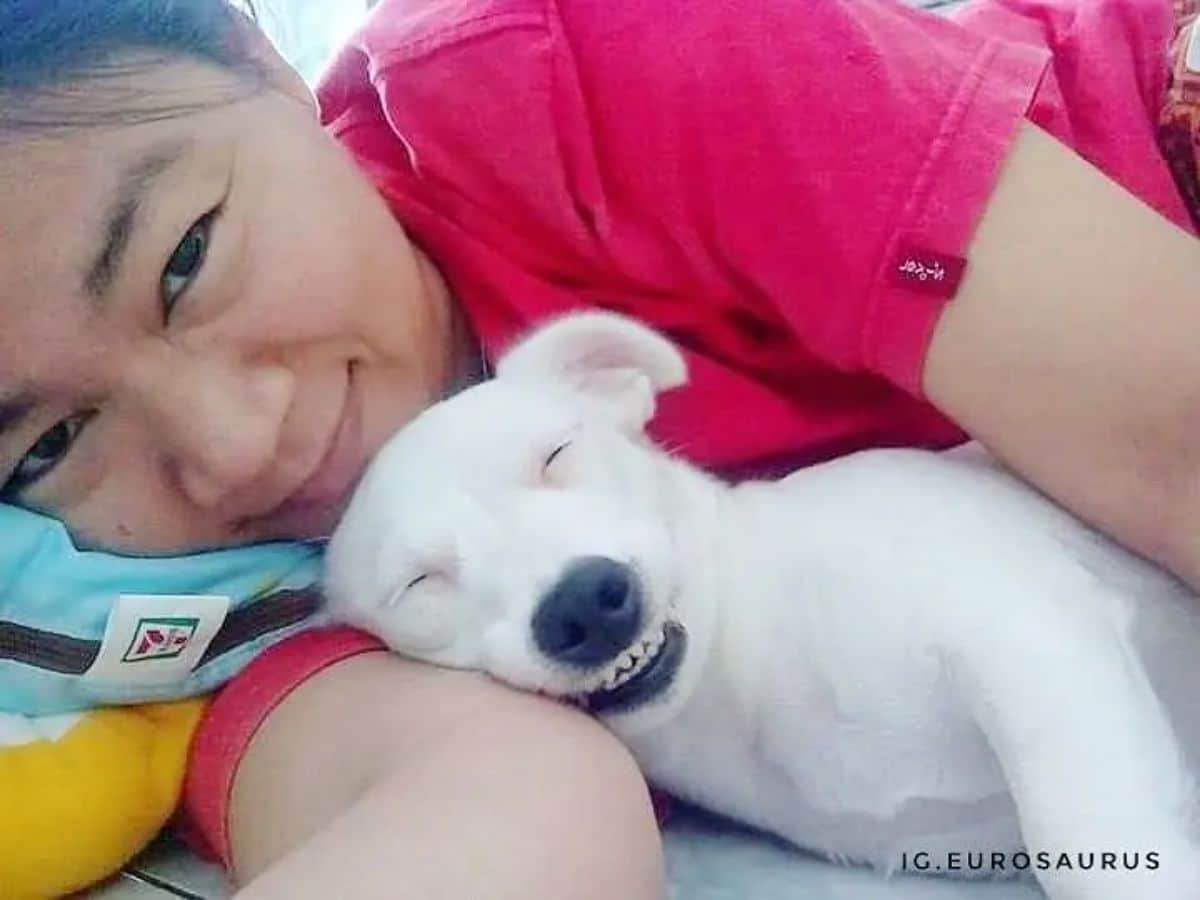 white dog cuddling sideways with a woman on a bed