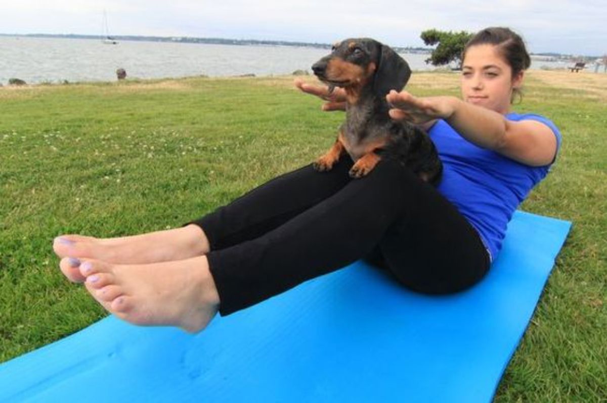 woman on yoga mat on grass with legs and arms lifted off the ground and stretched out with a black and brown dachund on her lap