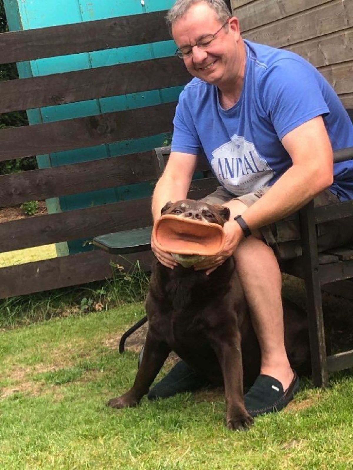 brown dog sitting by a man's feet with a large brown toy in the dog's mouth